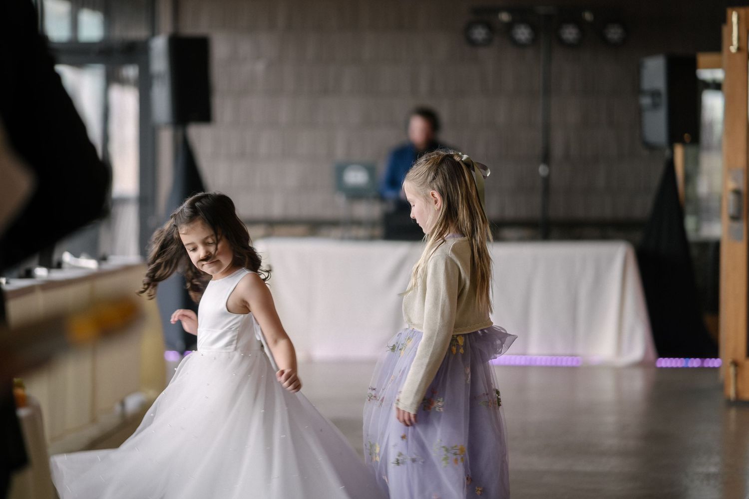 Two young children in flowing white dresses dance and twirl together on a dance floor at an event.