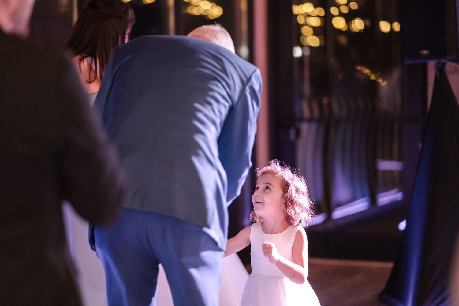 A small child in a white dress dances with an adult at a dimly lit evening reception.