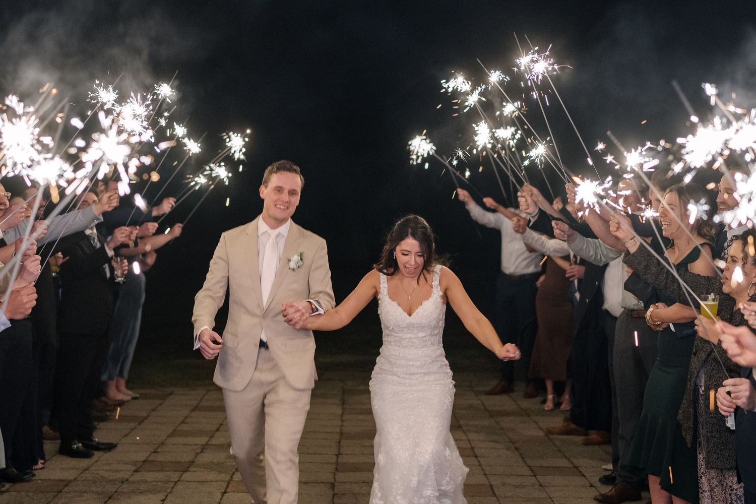 Wedding couple walks through sparkler exit tunnel with guests holding lit sparklers during nighttime celebration.
