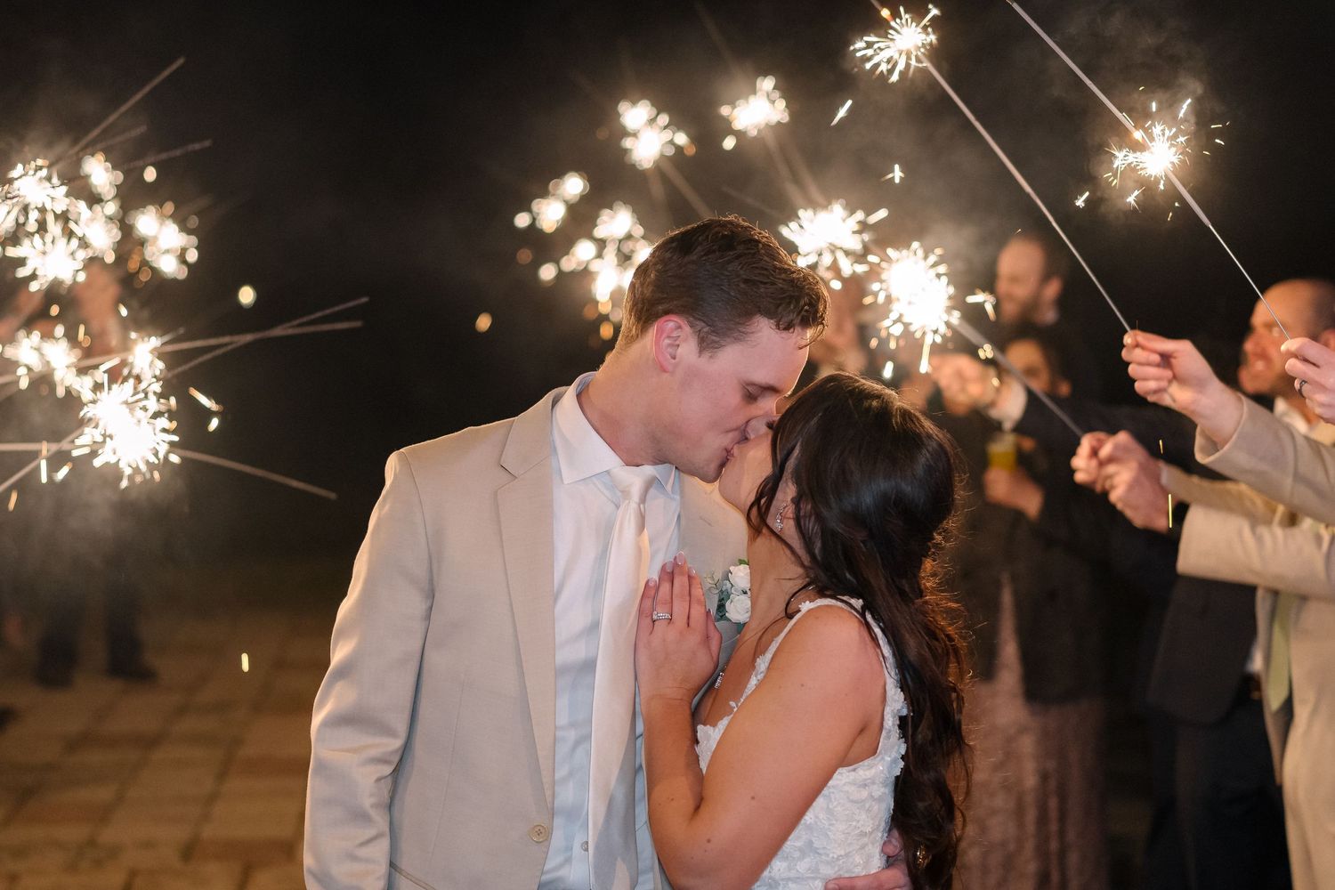 A couple shares an intimate moment while wedding guests hold sparklers during an outdoor nighttime celebration.