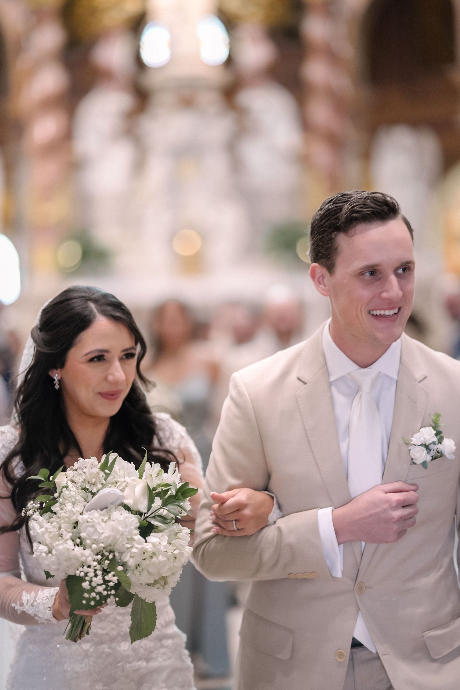 A wedding couple walks arm in arm at their ceremony, with the bride holding a white floral bouquet.