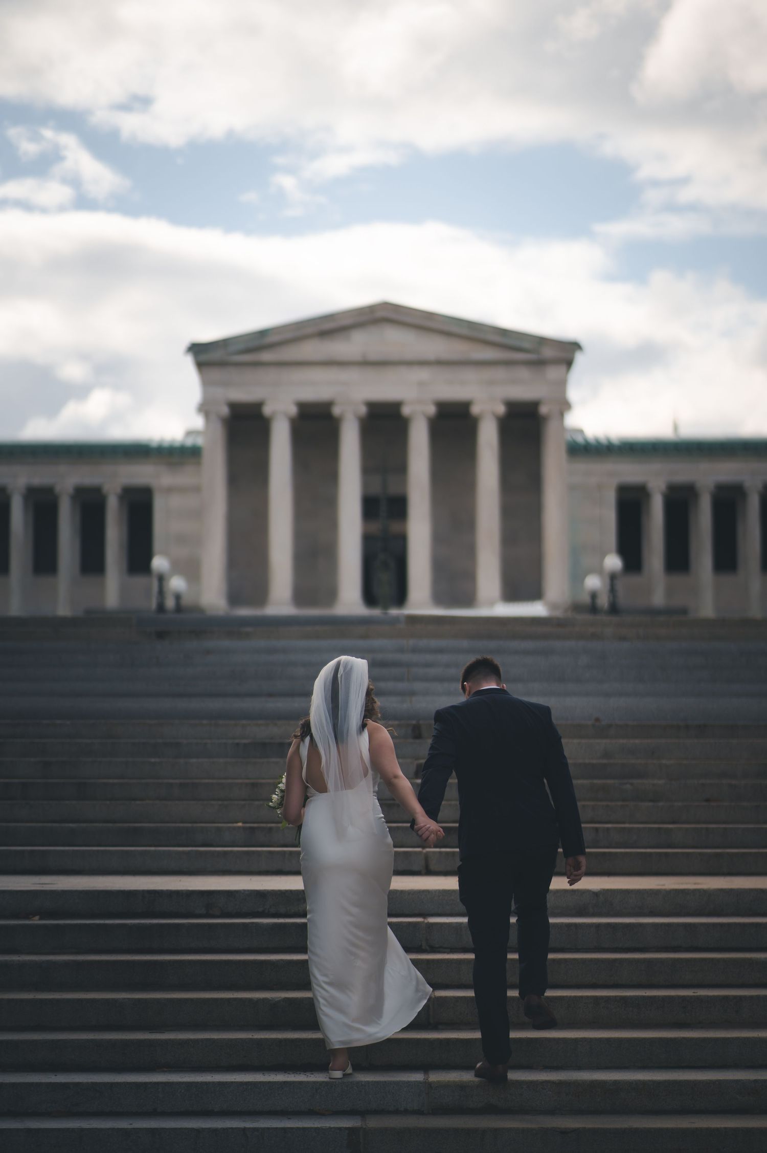 A newlywed couple ascending stone steps towards a neoclassical building with columns.
