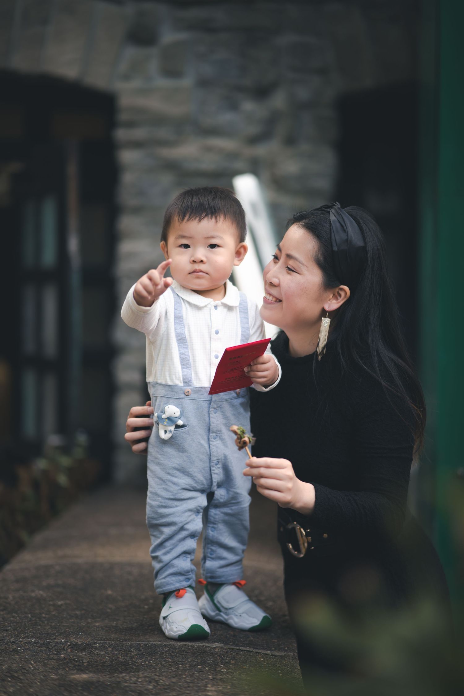A young child in denim overalls standing with a caregiver in black outdoors.
