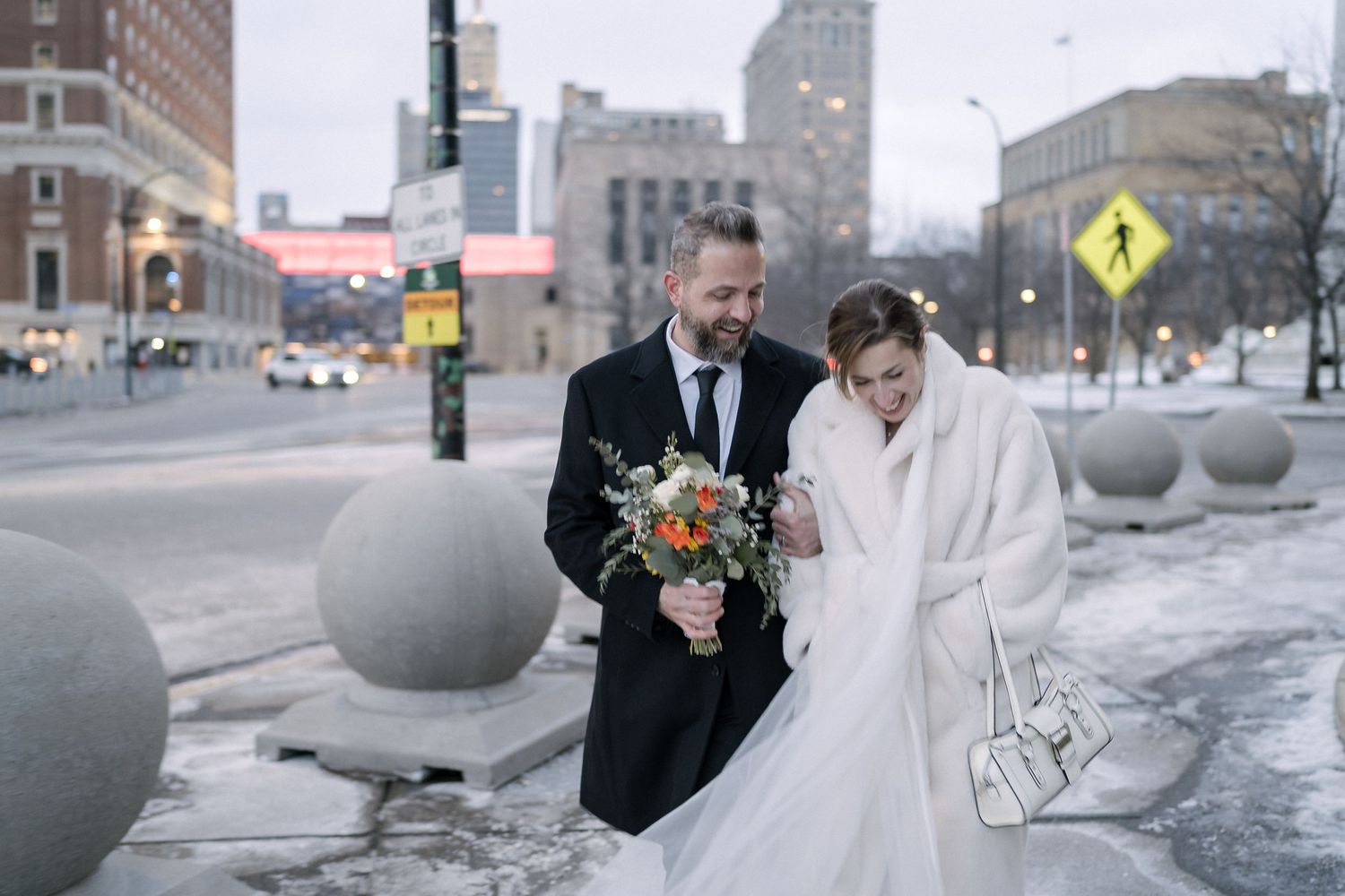 A winter wedding couple walks together through a snowy city street with decorative concrete bollards. Buffalo City Hall