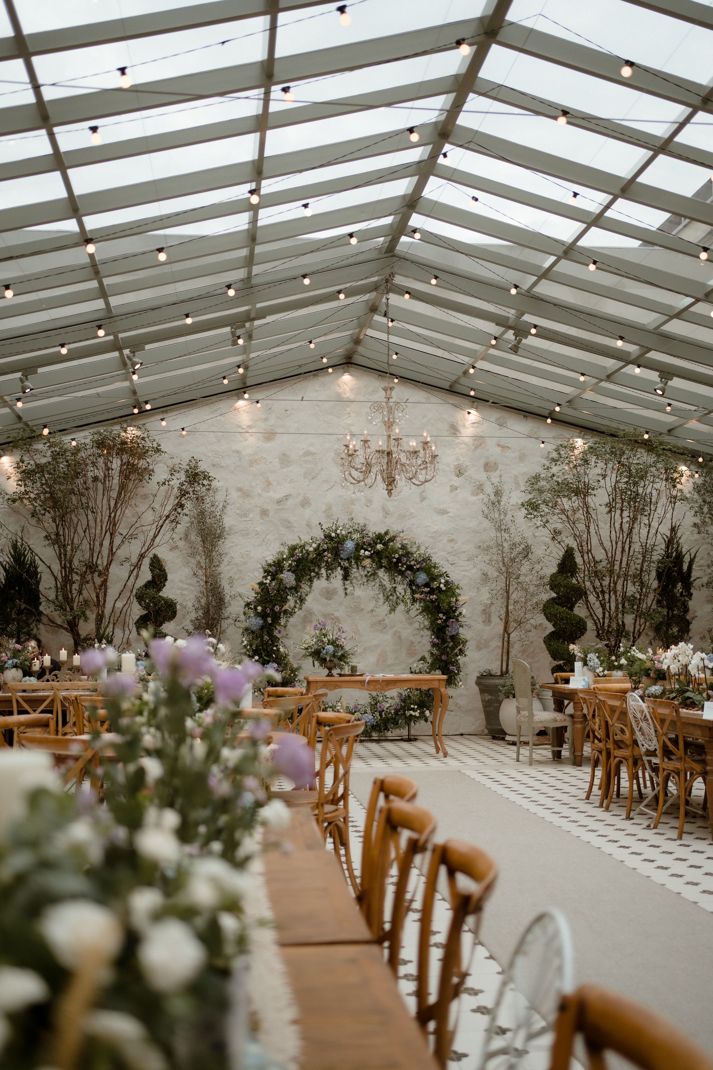 Wedding reception venue with glass ceiling, string lights and rustic wooden chairs in an elegant setup.