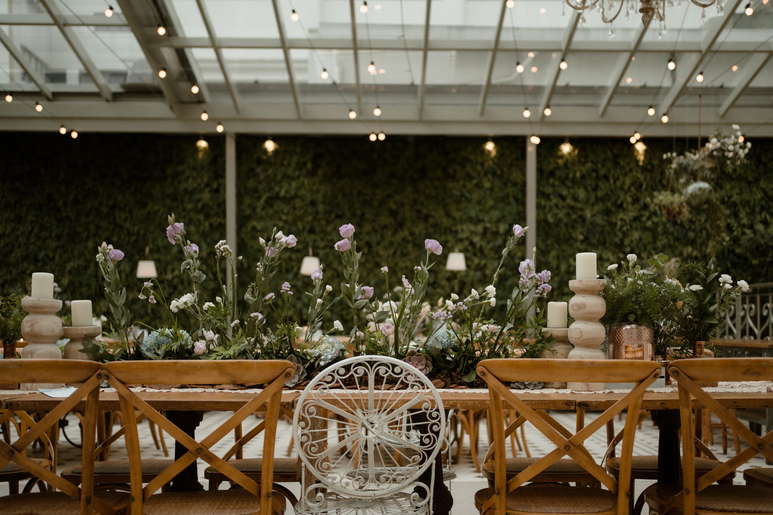 Elegant wedding reception venue with wooden crossback chairs and glass ceiling showcasing natural light and greenery.