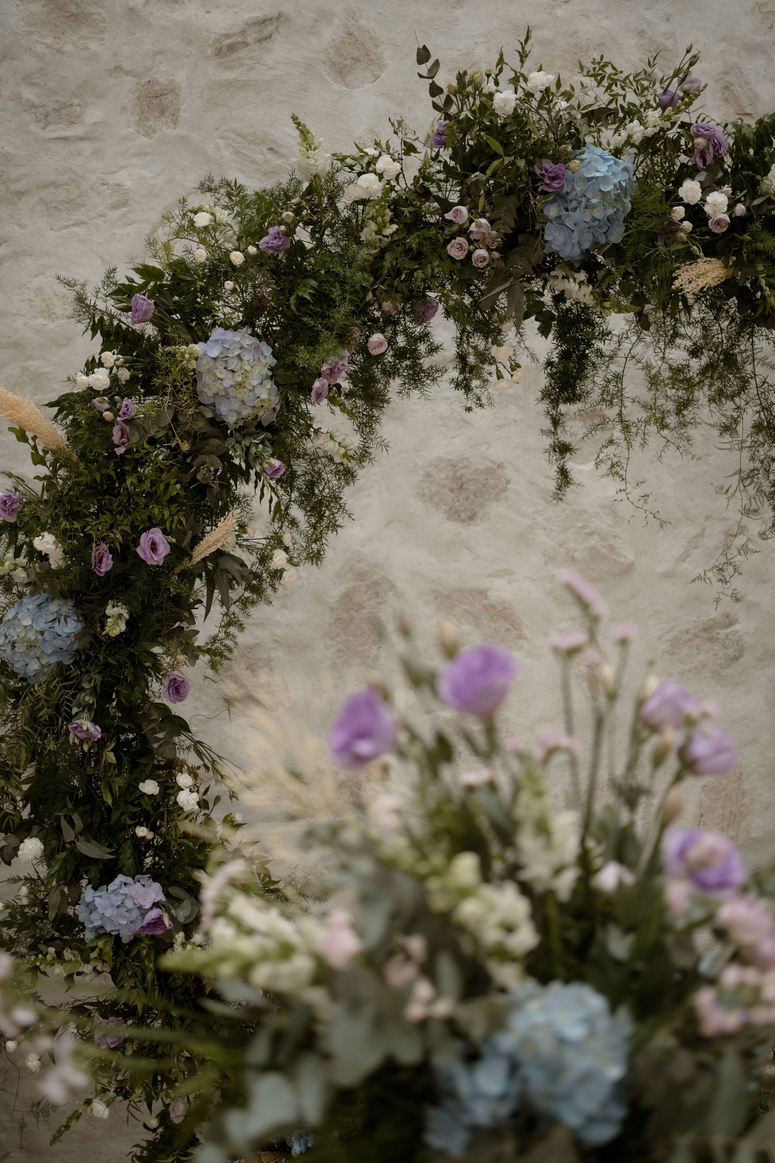 Floral arch with cascading white, pink, and blue flowers against a textured wall.