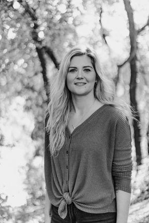 Black and white portrait of smiling blonde woman in tied knit cardigan standing among bokeh trees.