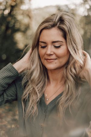 Blonde woman in olive green cardigan with eyes closed, gently touching her hair in autumn setting.