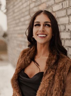 Smiling woman with dark hair wearing a brown fur coat and black dress against a brick wall.