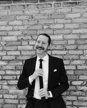 Laughing man in black suit adjusting his tie against a brick wall in black and white photo.