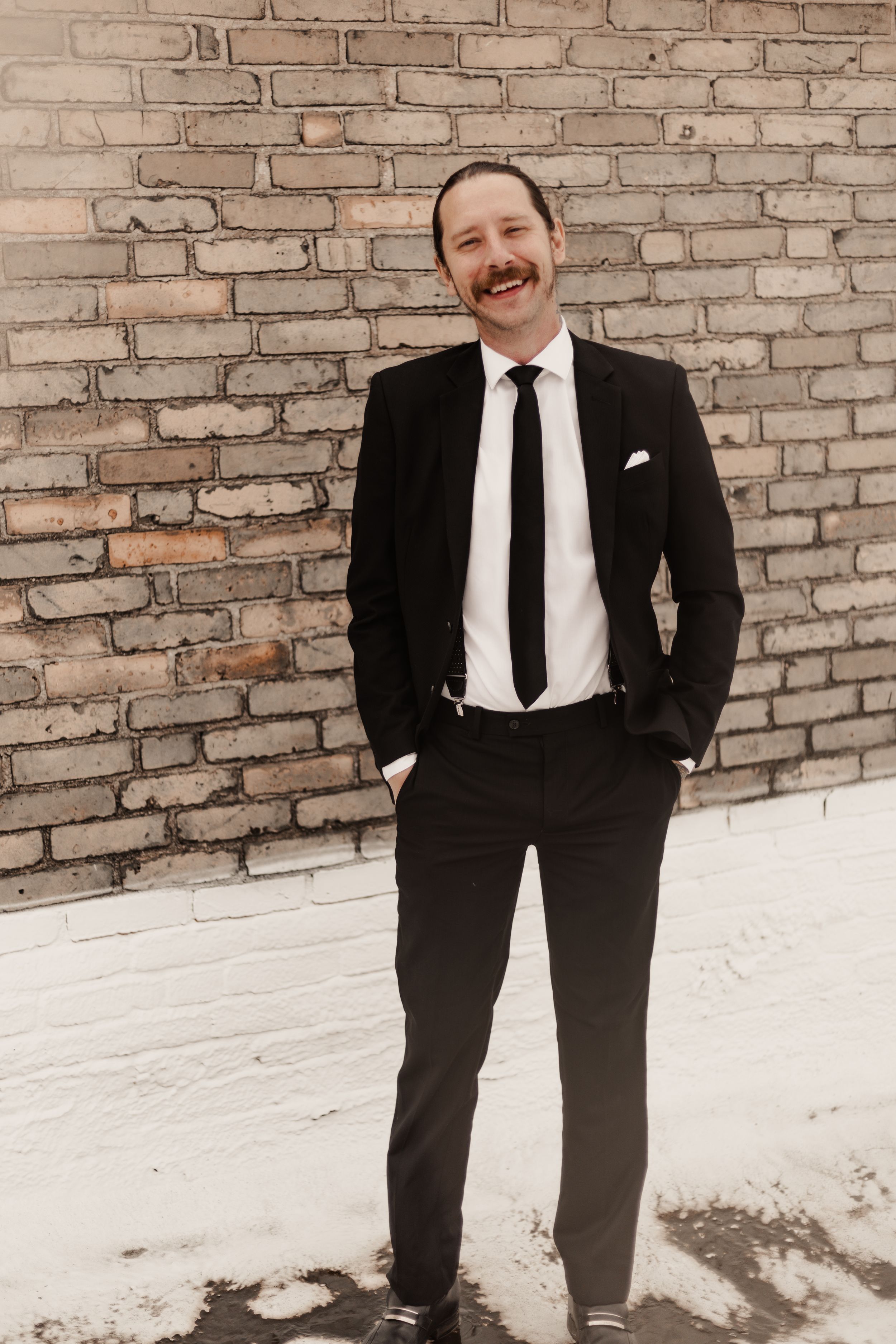Smiling man with mustache in black suit standing outdoors against a brick wall in winter.