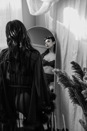 Black and white boudoir photo of woman in lingerie posing before a round mirror.