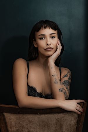 Young woman with tattoos and dark hair poses thoughtfully in black lace lingerie against a deep teal background.