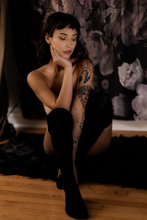 Tattooed woman in black bodysuit and knee-high socks poses gracefully against a floral backdrop on hardwood floor.