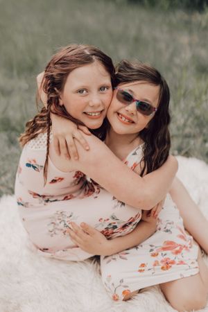 Two young sisters in floral dresses hugging and smiling on a white fur blanket outside.
