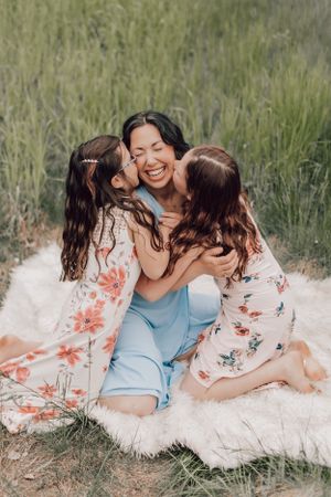 Mother laughing joyfully as two daughters kiss her cheeks while sitting on a blanket outdoors.