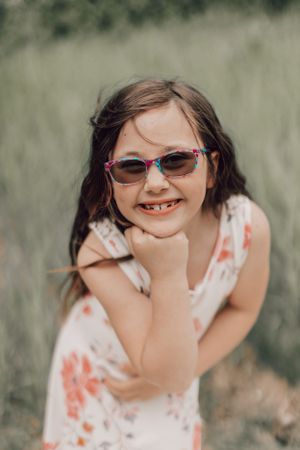 Young girl in sunglasses and floral dress smiling playfully with chin resting on hand outdoors.