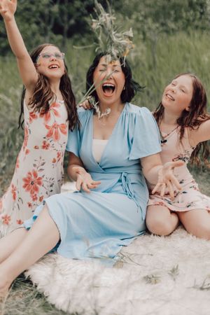 Mother and two daughters laughing and tossing wildflowers on a white blanket in a meadow.