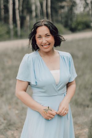 Smiling woman in a light blue wrap dress standing outdoors in a natural wooded setting.