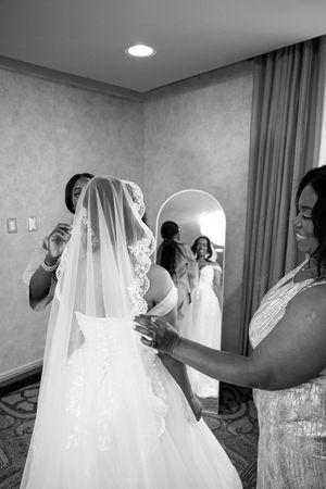 Black and white photo of bridesmaids helping a bride with her veil and wedding gown in a hotel room mirror.