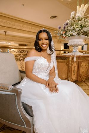 Smiling bride in an off-shoulder lace ballgown seated in an ornate venue chair with floral arrangement nearby.