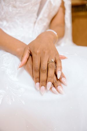 Close-up of a bride's hands with elegant almond-shaped nails and a diamond engagement ring on white dress.