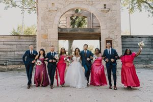 Wedding party in navy suits and pink gowns walking together under a stone arch at outdoor venue.