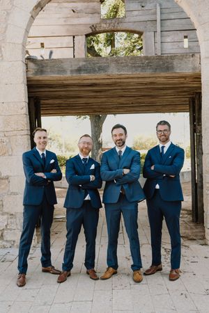 Four groomsmen in matching navy suits with brown shoes posing with crossed arms under a rustic wooden arch.