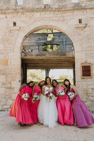 Bride with four bridesmaids in mixed pink gowns holding bouquets under a rustic stone archway.