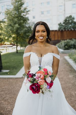 Radiant bride in off-shoulder lace ballgown holding a colorful pink and red bouquet in an outdoor garden.
