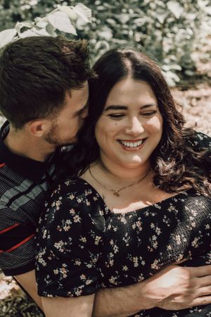 Man kissing smiling woman on cheek outdoors, woman in black floral dress laughing joyfully.