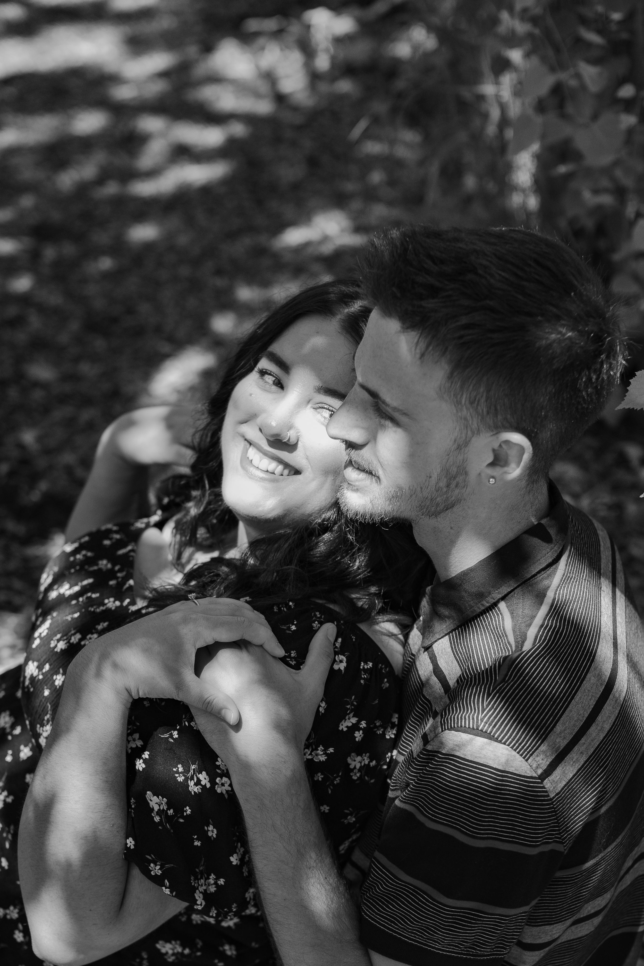 Black and white photo of a smiling couple embracing outdoors under dappled sunlight.