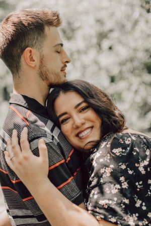 A happy couple hugs closely as the man kisses the womans head near blooming trees.
