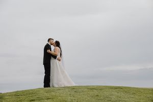 Bride and groom sharing romantic kiss on green hilltop under dramatic cloudy sky on wedding day.