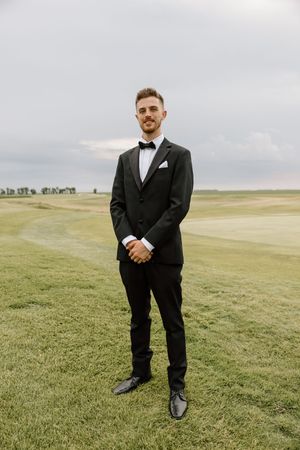 Groom in black tuxedo with bow tie standing confidently on green golf course under cloudy sky.