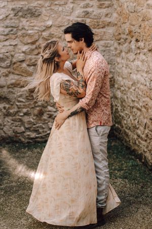 Couple embracing and smiling at each other near stone wall, woman in floral dress with arm tattoos.
