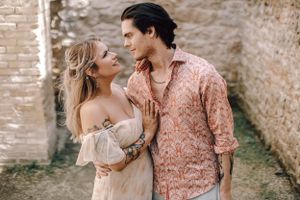 Couple gazing at each other near rustic stone wall, woman in white dress with tattoos and man in pink shirt.