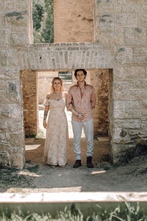 Bride and groom holding hands walking through rustic stone archway outdoors.