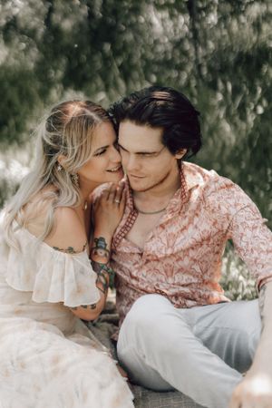 Tattooed blonde bride whispering to groom sitting together under green trees.