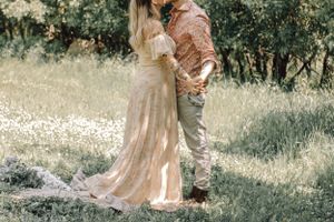 Couple embracing in sunlit meadow, woman in elegant floral gown and man in patterned shirt.