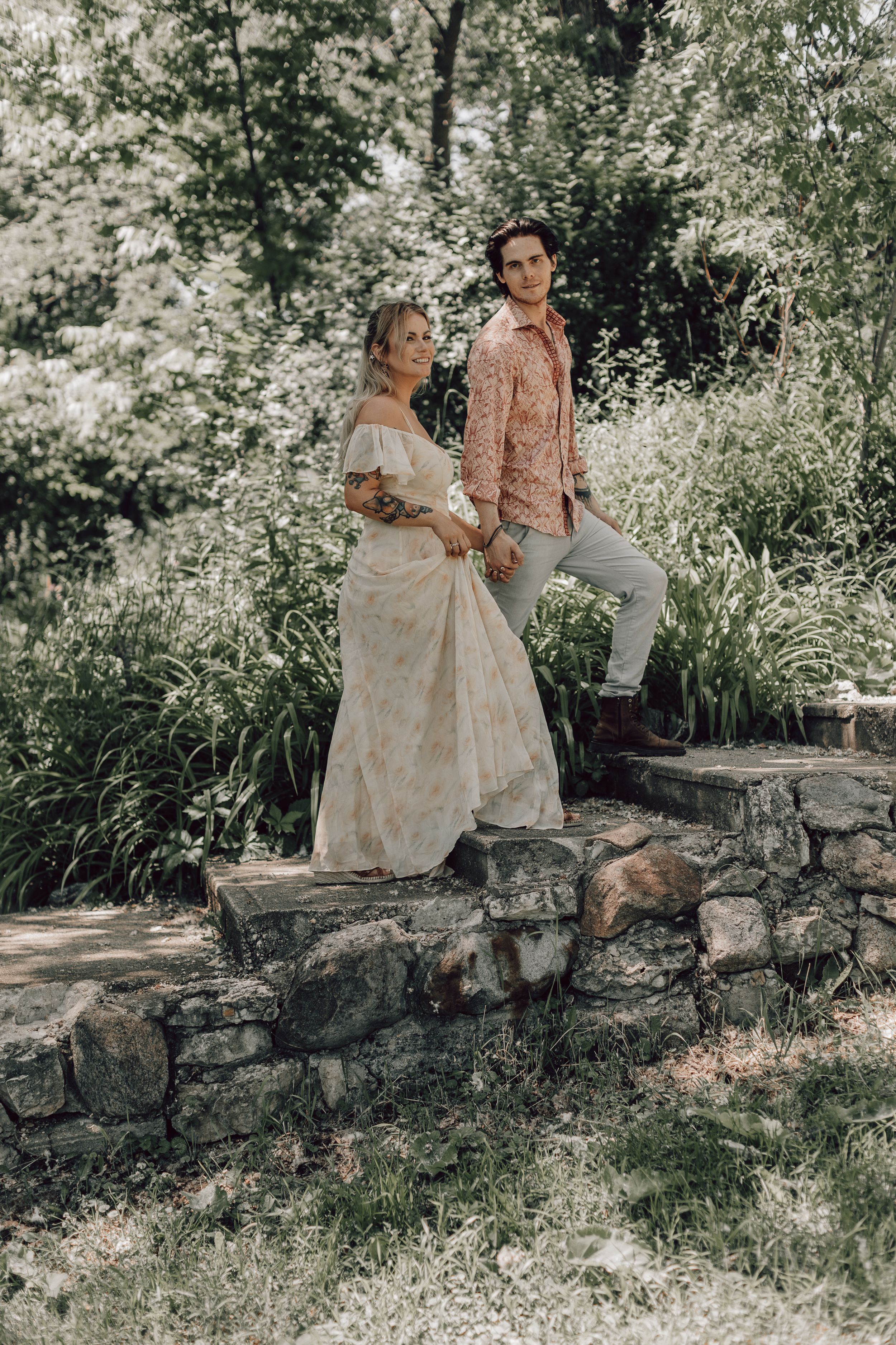 Couple posing on stone steps surrounded by lush greenery, woman in boho floral wedding dress.