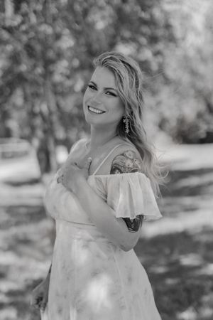 Black and white portrait of smiling blonde woman with tattoos in off-shoulder dress outdoors.