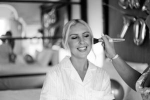 Smiling blonde woman getting makeup applied in black and white bridal preparation photo.