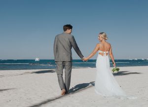 Bride and groom walking hand in hand on a white sand beach on a sunny day.