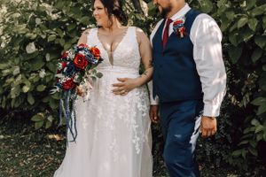 Bride in lace gown and groom in navy vest walking together holding a red and blue floral bouquet.