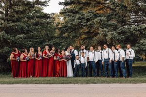 Large wedding party posing outdoors with bridesmaids in red dresses and groomsmen in navy suspenders.
