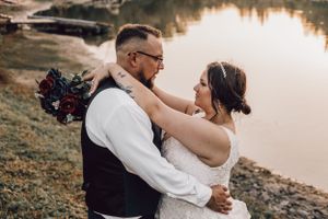 Bride and groom sharing a romantic moment by a lakeside, bride holding a deep red and navy bouquet.