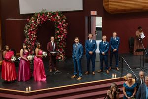 Wedding ceremony on stage with bridesmaids in red and pink gowns, groomsmen in navy suits, and a floral arch backdrop.