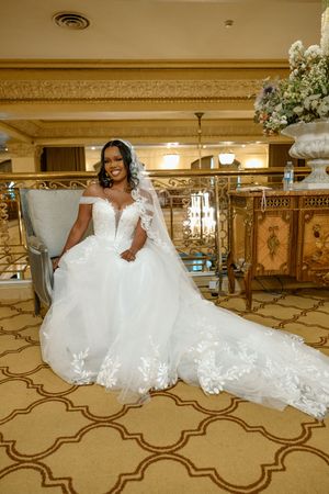 Radiant bride in elegant off-shoulder ball gown with long veil posing in ornate venue interior.